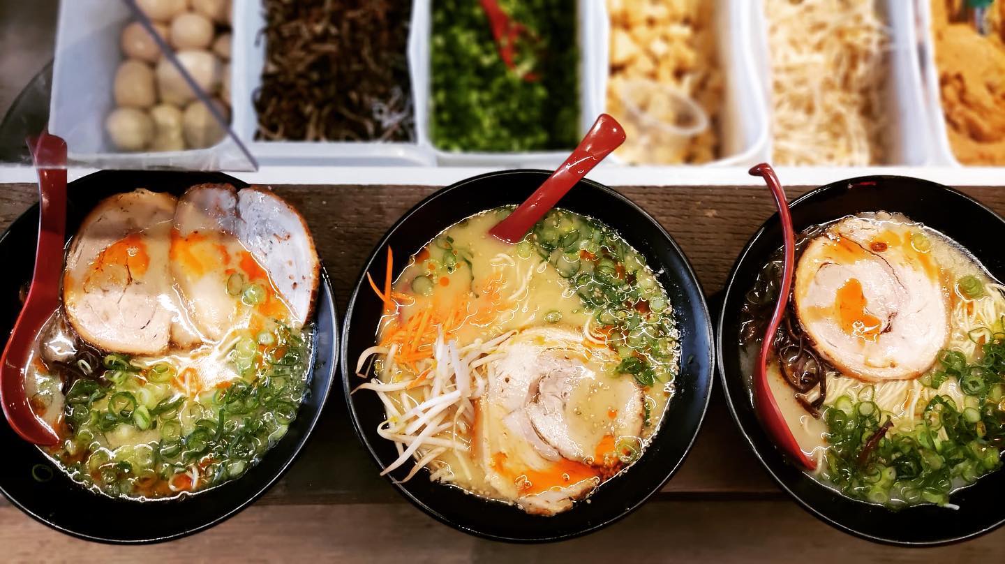 Three bowls of ramen with soft-boiled eggs, chashu pork, and scallions served in black bowls with red spoons.