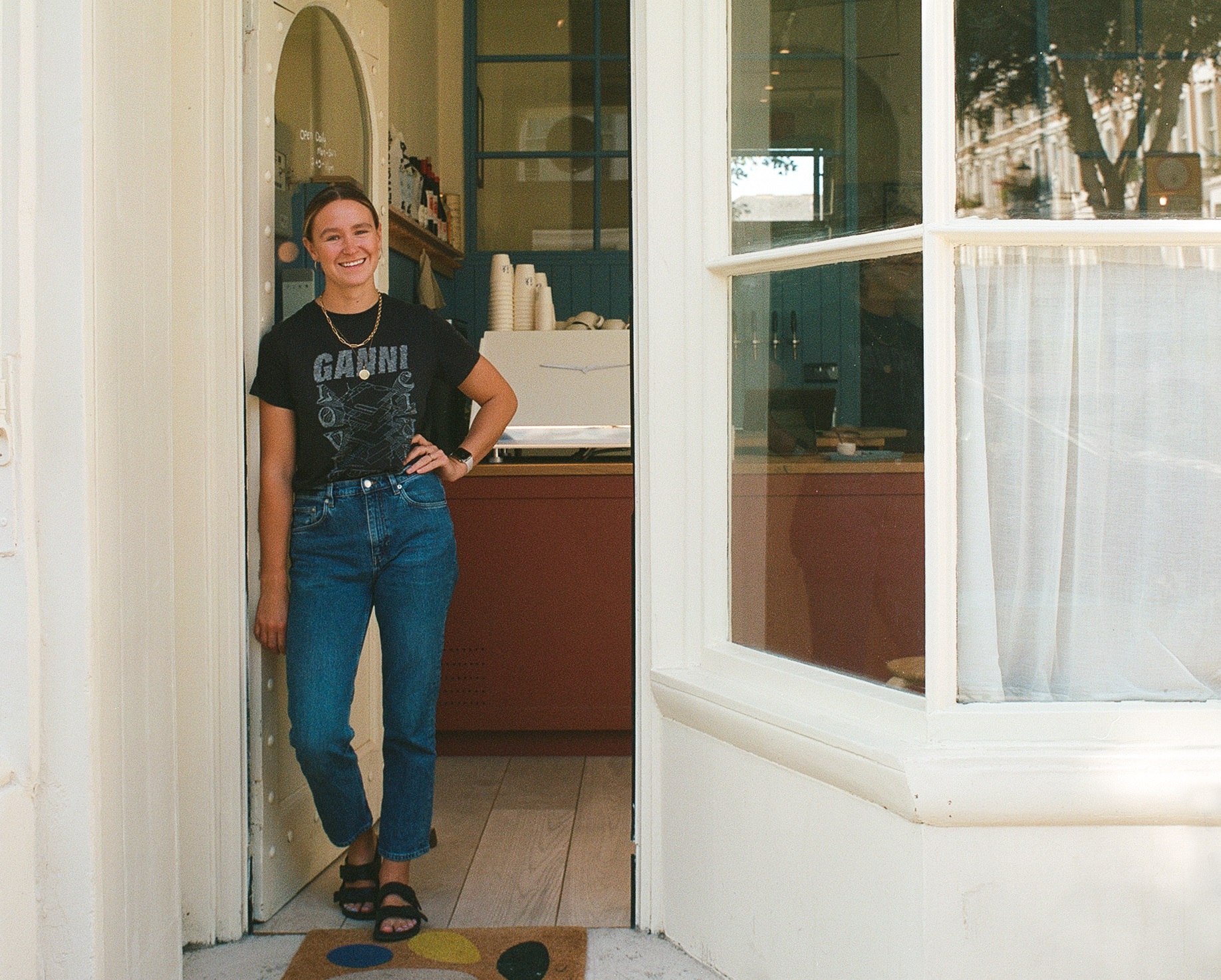 A smiling woman in jeans and a black t-shirt stands in the doorway of a bright, modern restaurant with white storefront windows.
