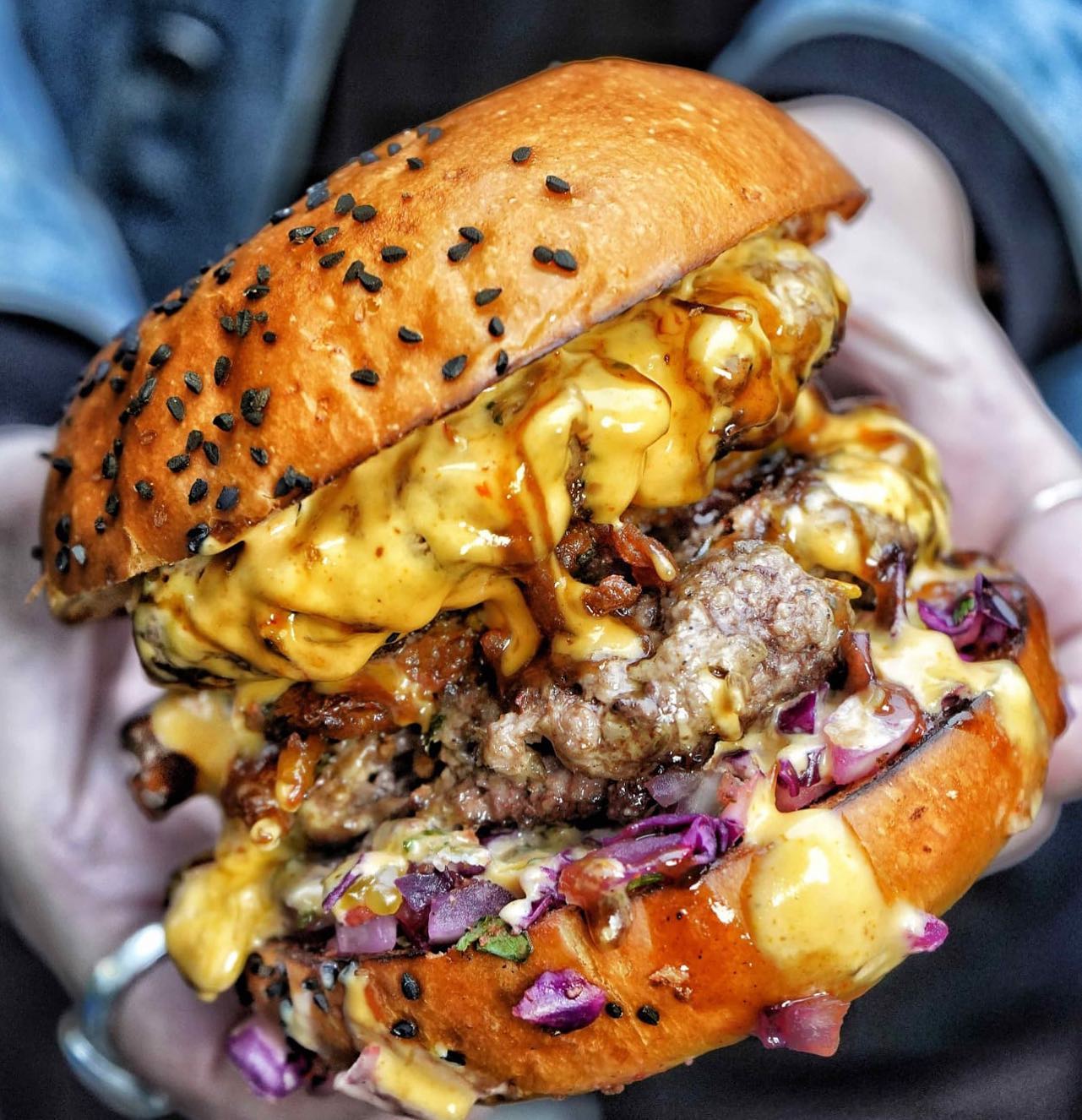 Hands holding a loaded burger with beef patty, melted cheese, caramelized onions, and purple cabbage on a sesame seed bun.