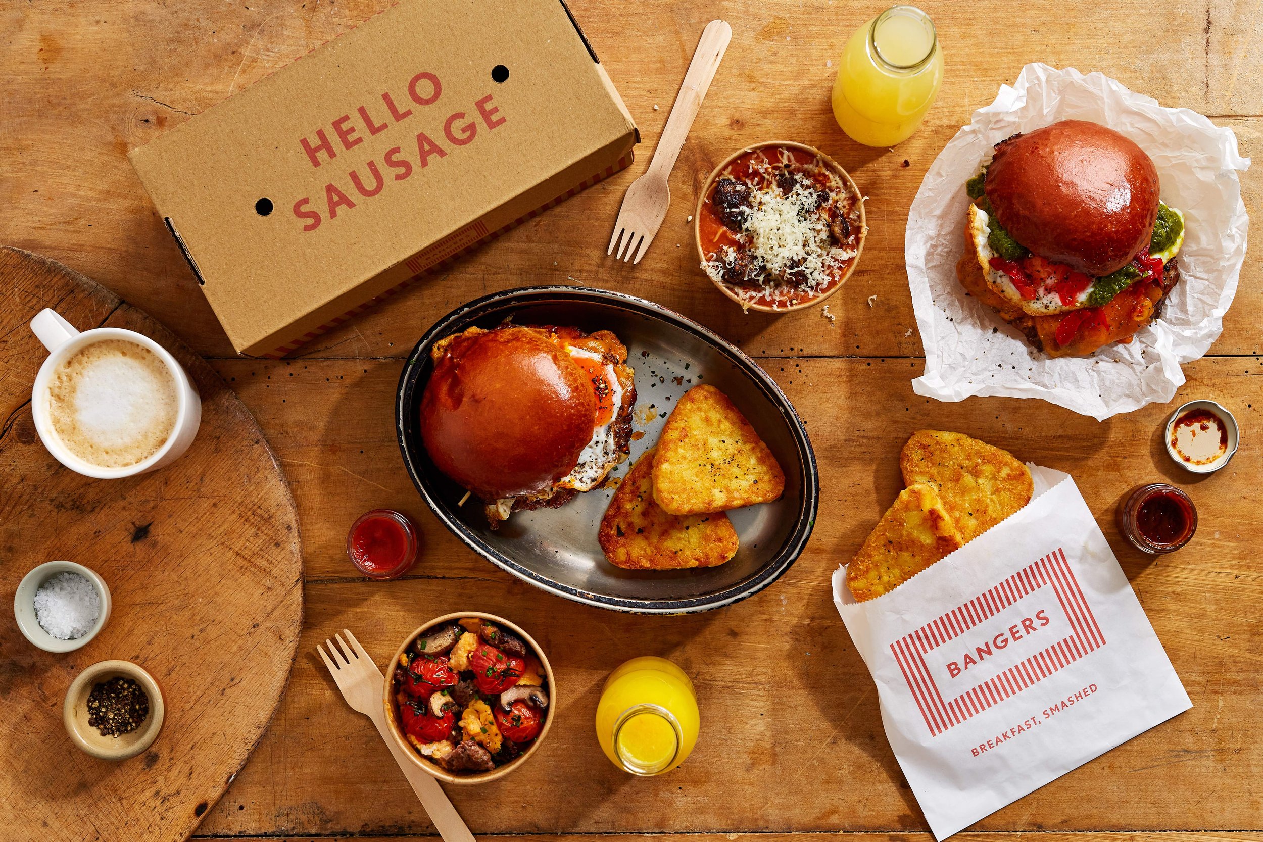 Gourmet sausage sandwiches with golden hash browns and sides spread across a wooden table at Bangers breakfast bar.