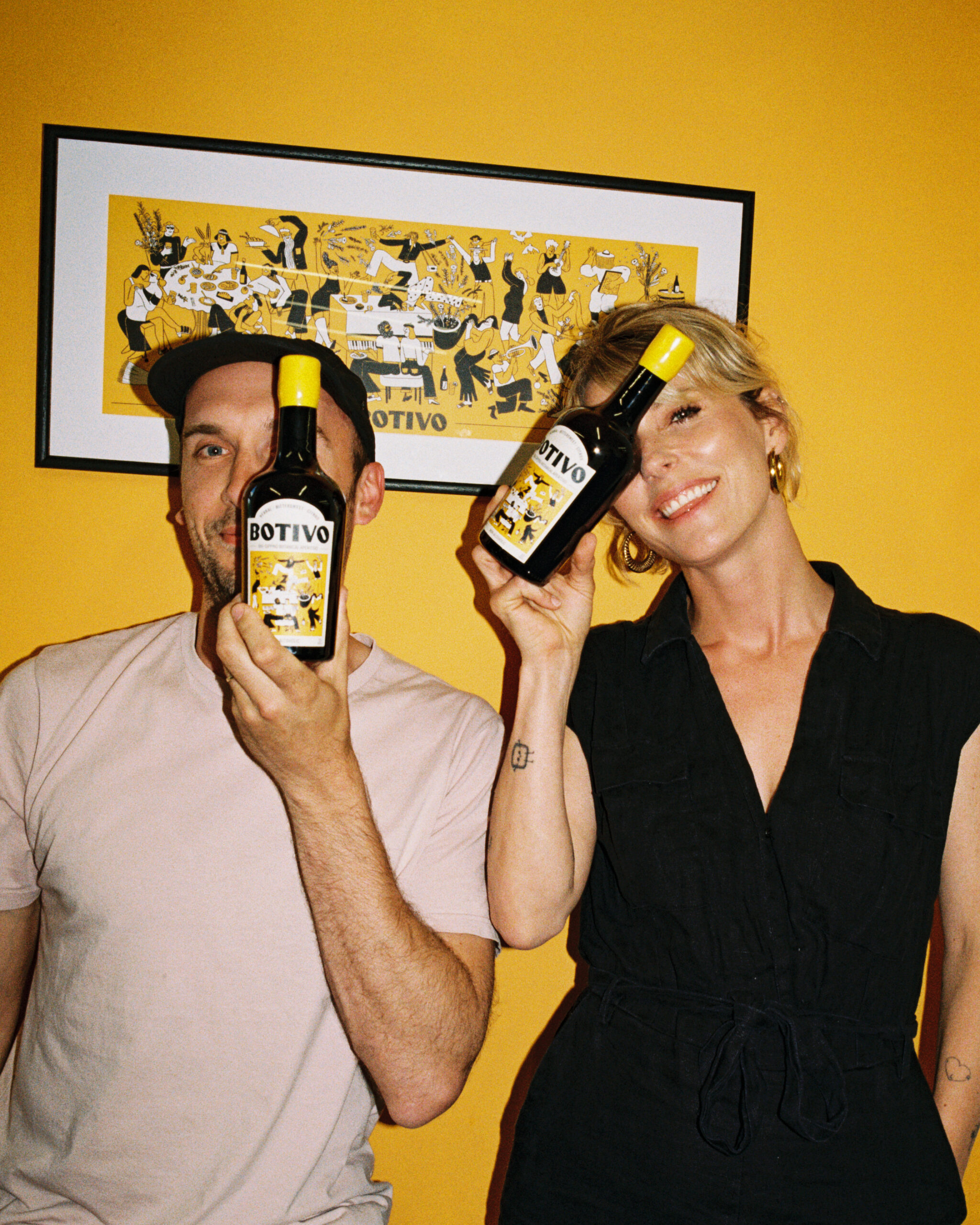 Two people playfully hold Botivo botanical aperitivo bottles up to their faces, smiling against a bright yellow wall.