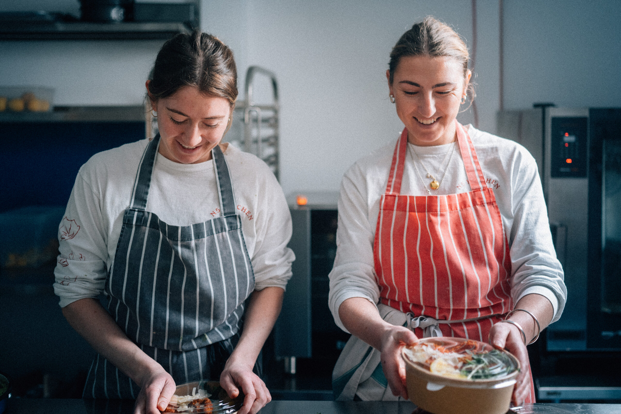 Two women in striped aprons smile while preparing food in a modern kitchen, one plating a dish and the other holding a bowl.