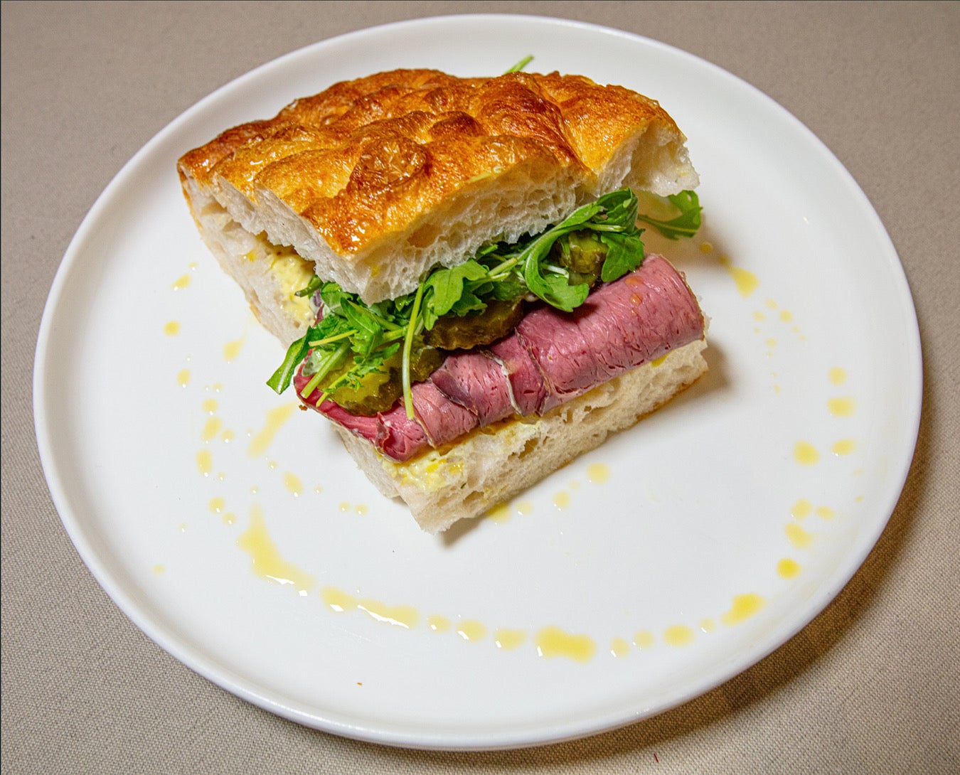 A gourmet sandwich with sliced roast beef, greens, and pickles on crusty bread, plated with olive oil drizzle.