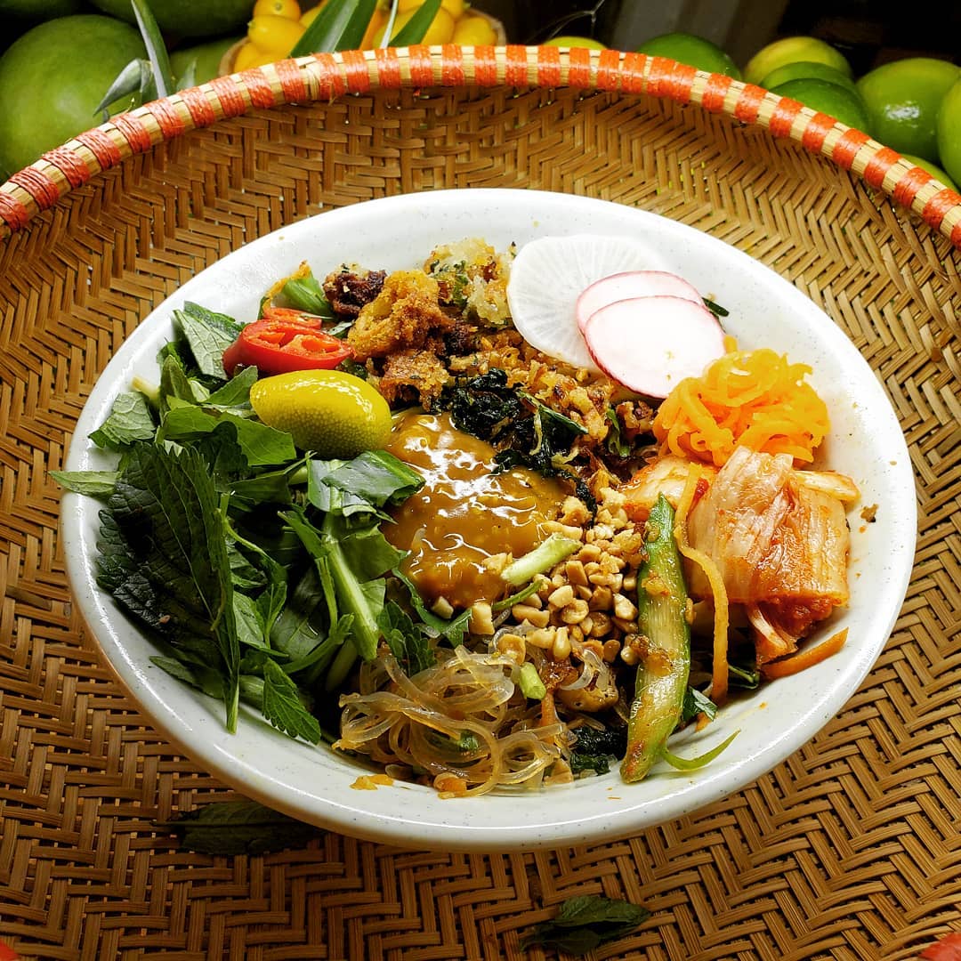A white bowl filled with Vietnamese bun thit nuong featuring grilled pork, fresh herbs, pickled vegetables, and rice noodles in a woven basket.