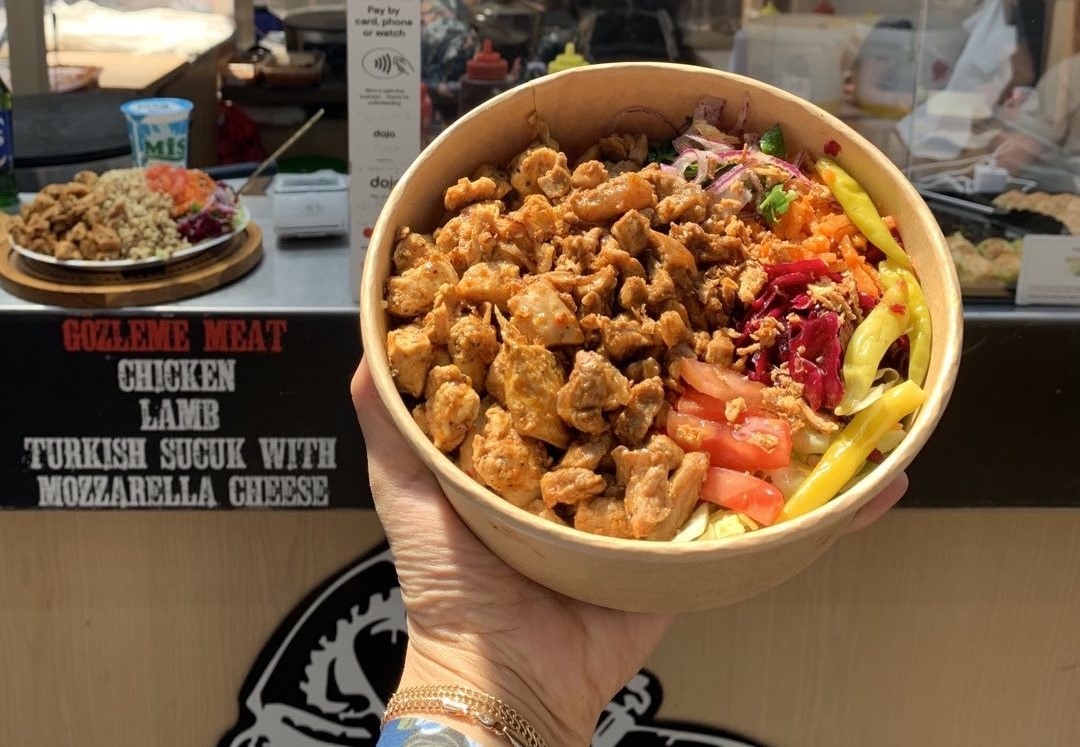Hand holding a bowl of Middle Eastern rice with spiced meat, pickled vegetables, and fresh herbs at a market food stall.