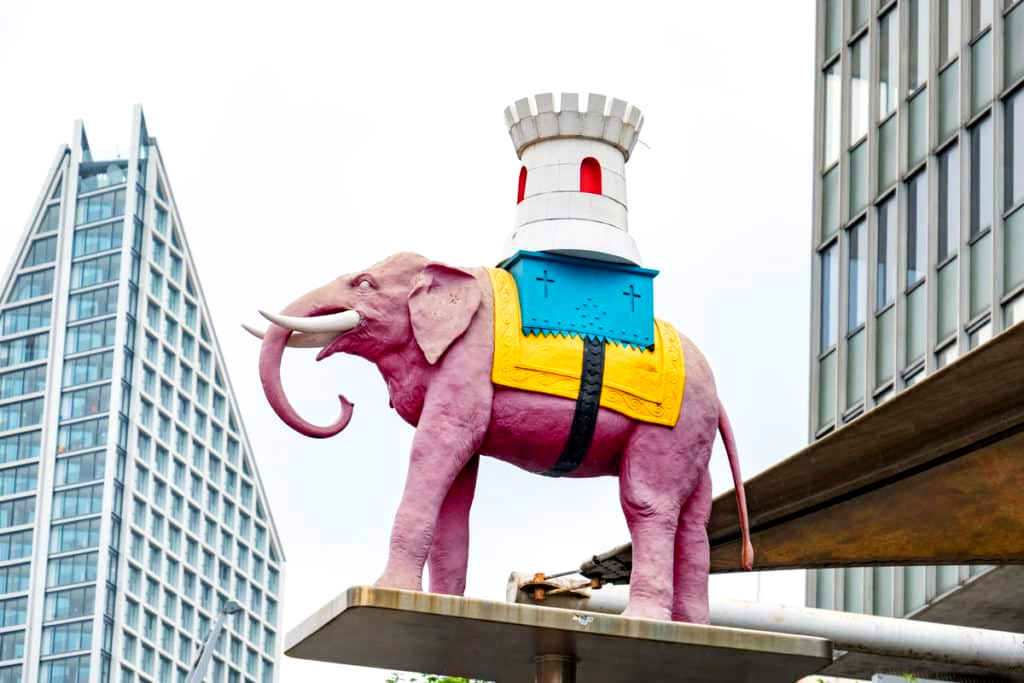 A pink elephant statue wearing a white castle tower on its back stands on a platform between modern buildings in an urban setting.