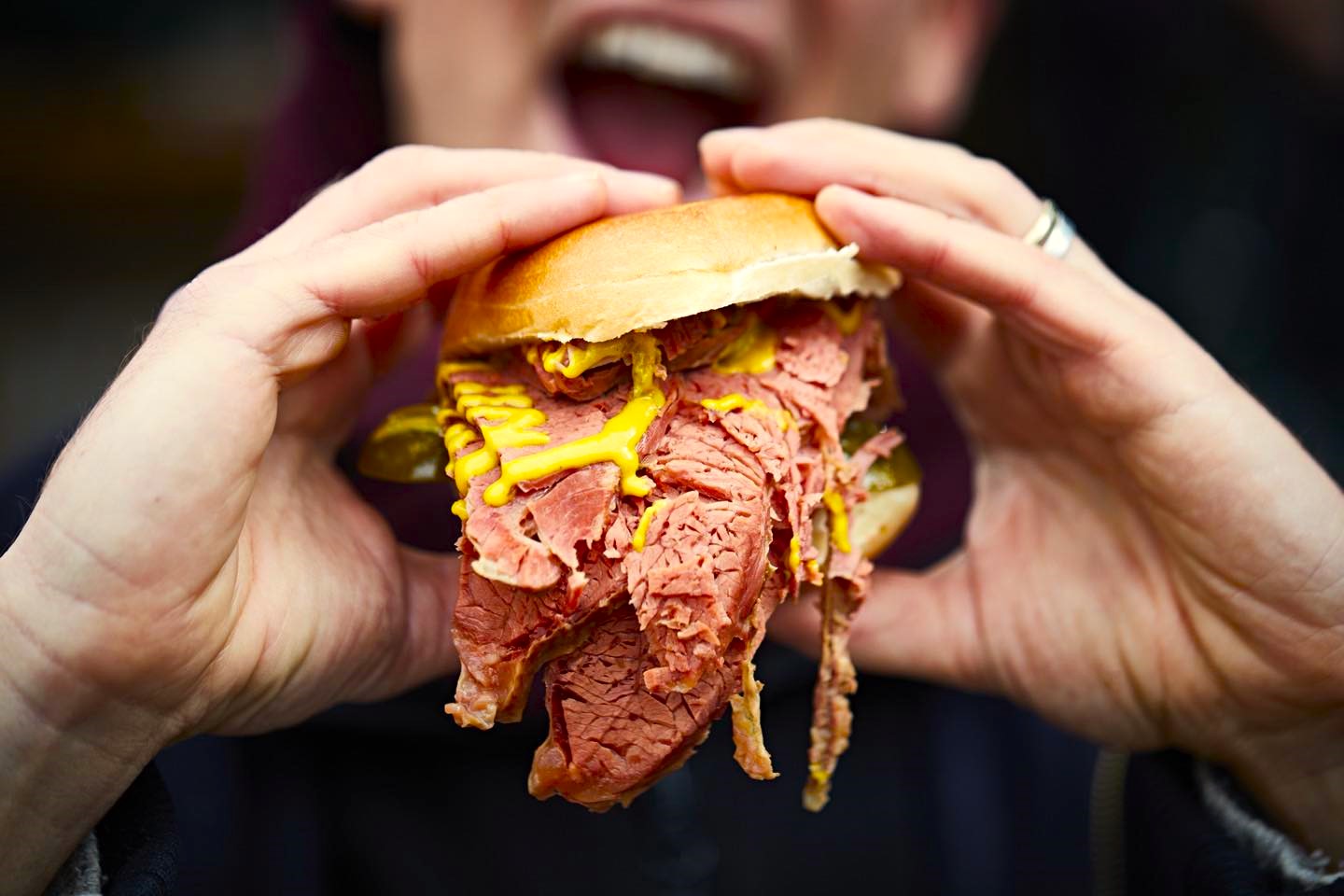 Hands holding an overstuffed deli sandwich with thick-cut pastrami and yellow mustard dripping from a crusty roll.