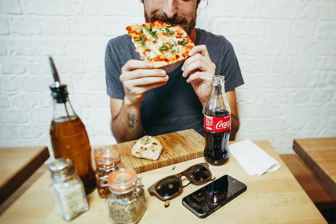 A bearded man in a gray t-shirt takes a bite of pizza with a Coca-Cola bottle on a wooden table beside sunglasses and phone.