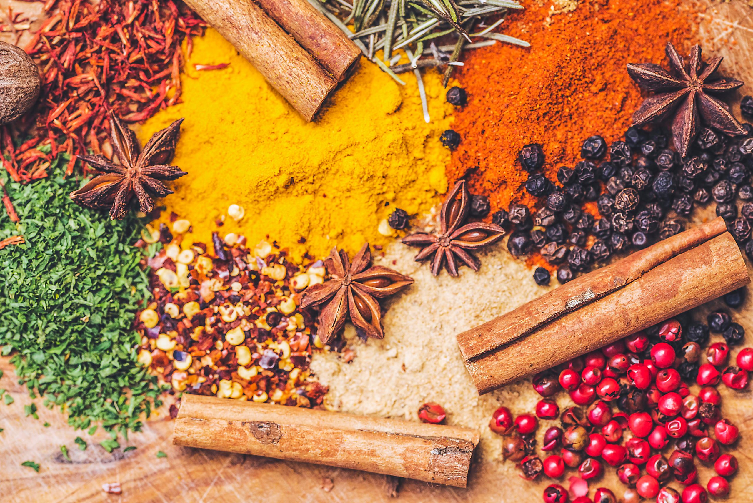Colorful array of whole spices including cinnamon sticks, star anise, turmeric, paprika, peppercorns, and dried herbs arranged on wood.