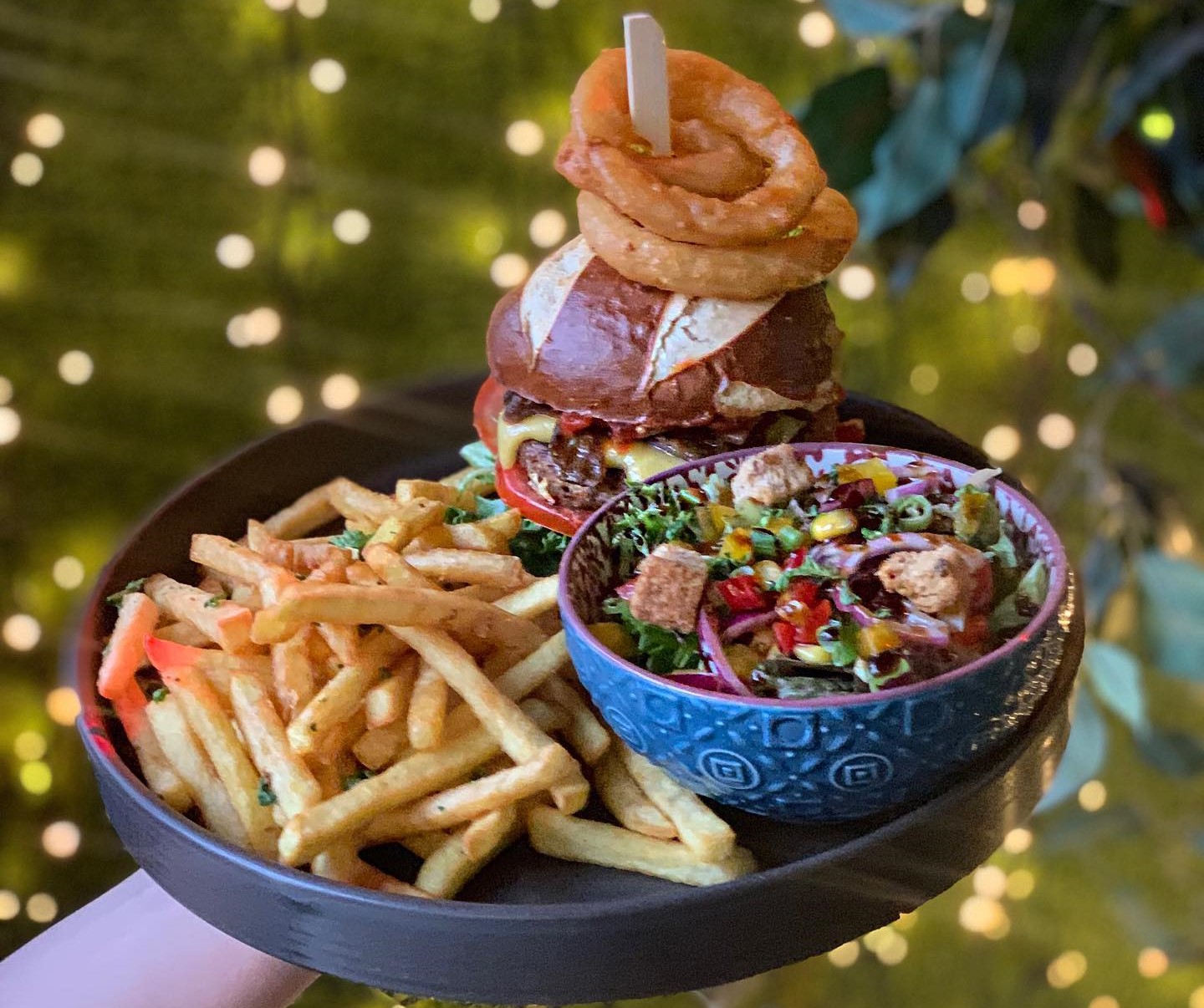 A gourmet burger with onion rings on top, served with french fries and a colorful salad bowl on a dark wooden platter.