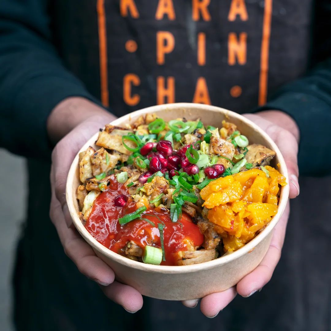 A person holds a wooden bowl filled with colorful grains, roasted vegetables, pomegranate seeds, and fresh herbs.