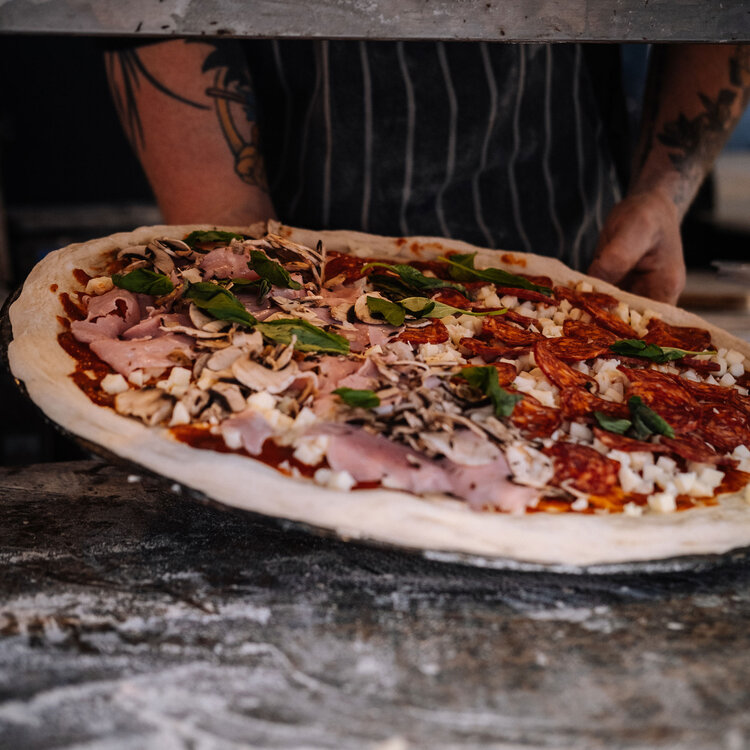 A pizza maker slides a freshly topped pizza with cured meat, mushrooms, and greens into a wood-fired oven at Breadstall.