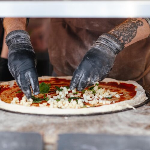 Pizza chef adding fresh cheese and herbs to an uncooked pizza with red sauce before placing it in a wood-fired oven.