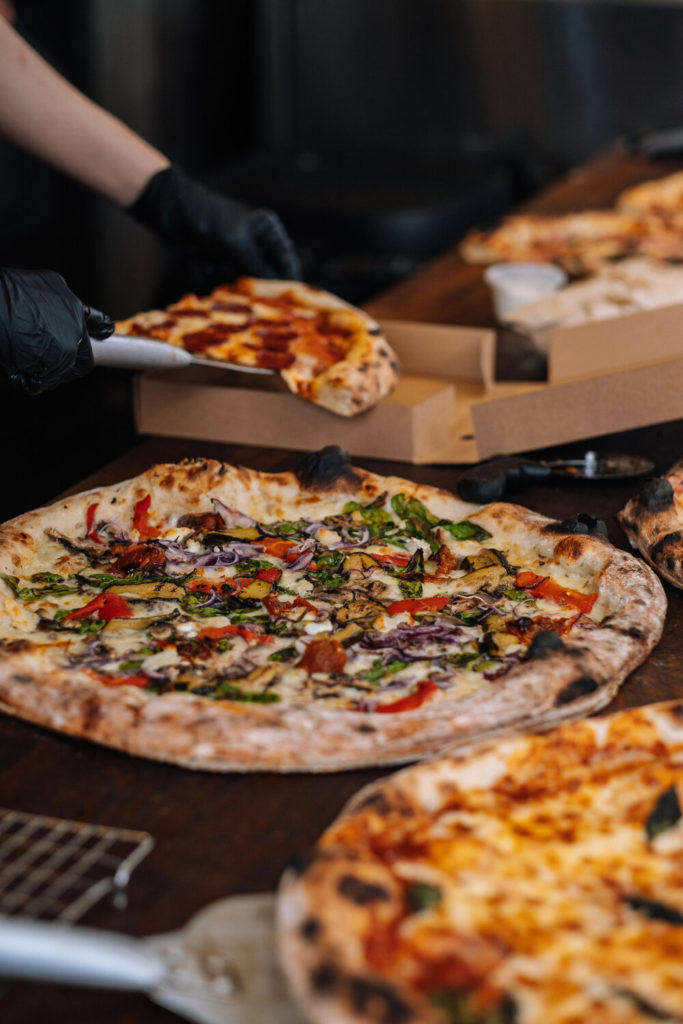 A pizza chef in black gloves places a slice into a takeaway box, with freshly baked pizzas topped with vegetables and cheese displayed on the counter.