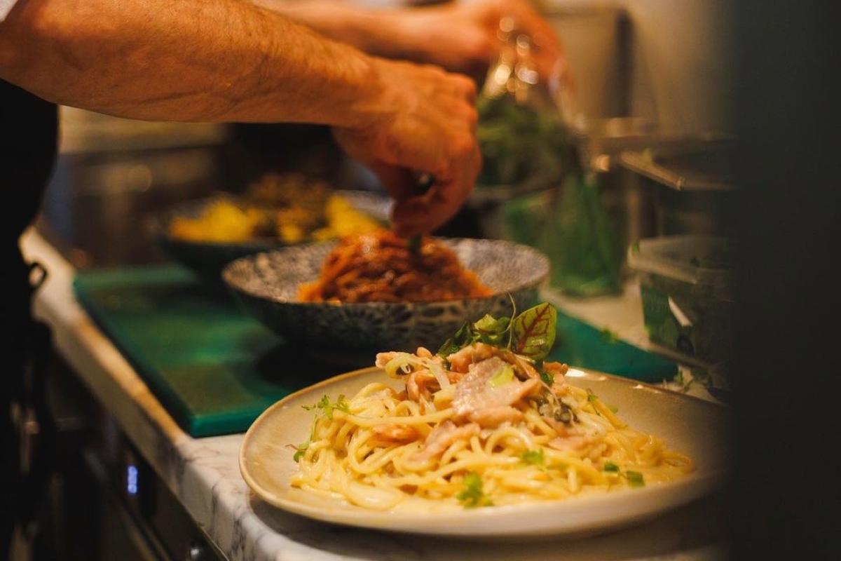 A chef's hands grate fresh cheese over a plate of pasta with herbs in a restaurant kitchen.
