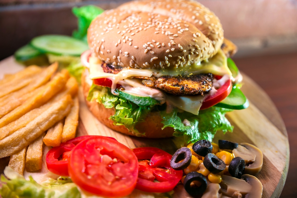 A plant-based burger with sesame seed bun, lettuce, tomato and cheese served with golden fries and sliced vegetables on a wooden board.