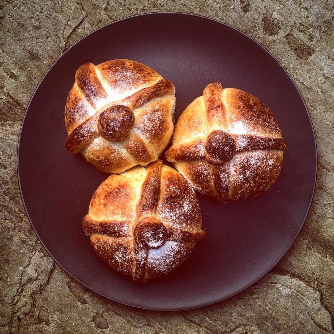 Three golden pan de muerto breads with cross patterns and circular decorations arranged on a dark purple plate.