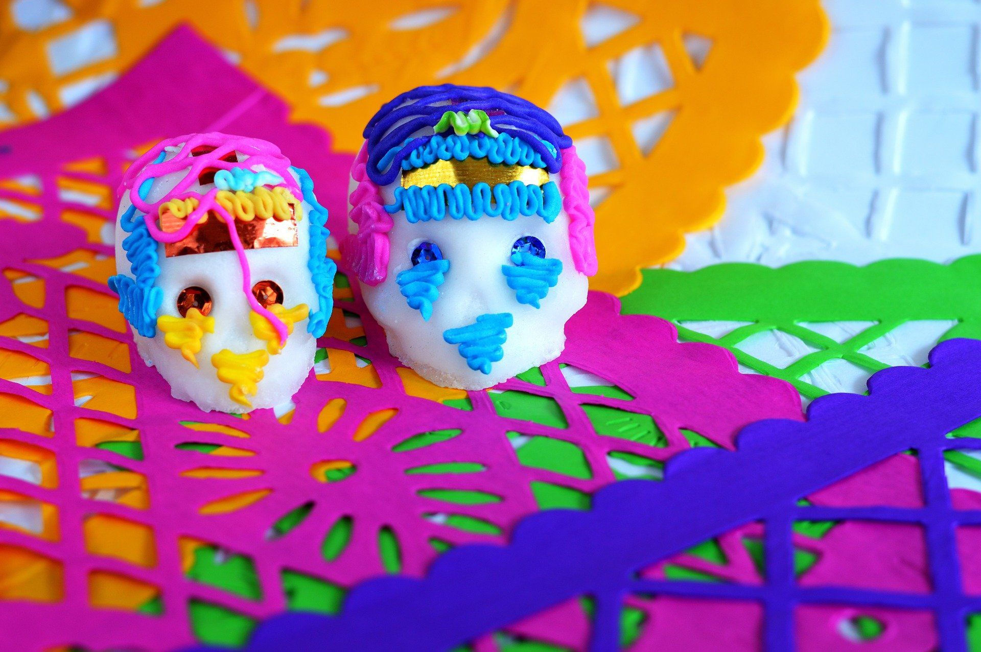 Two decorated sugar skulls with colorful icing sit among vibrant papel picado decorations in bright pink, orange, and green.