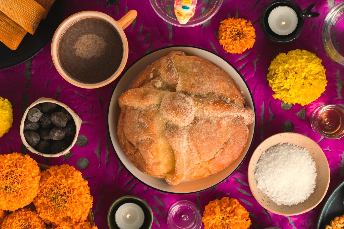 Traditional Día de Muertos bread (pan de muerto) surrounded by marigold flowers, candles, and ceremonial offerings on a vibrant purple backdrop.