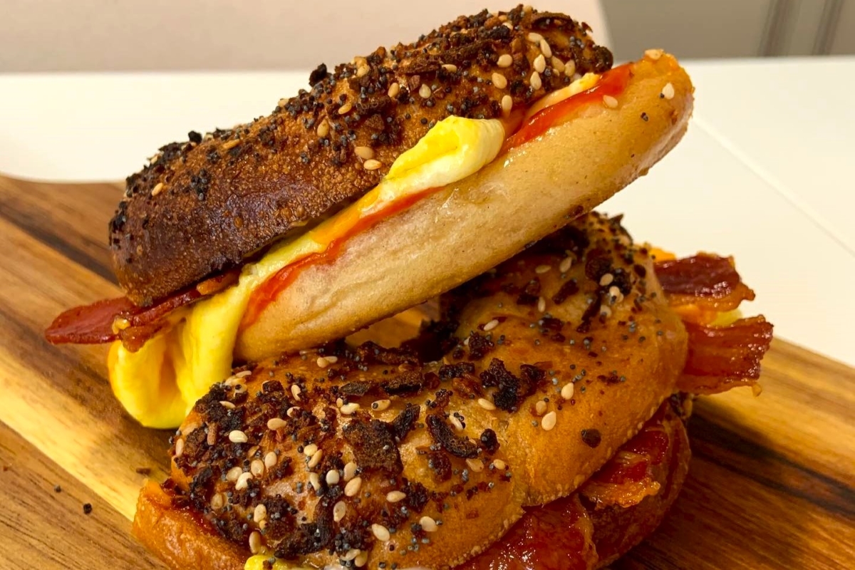 Everything bagels topped with mustard, bacon, and sesame seeds stacked on a wooden cutting board.