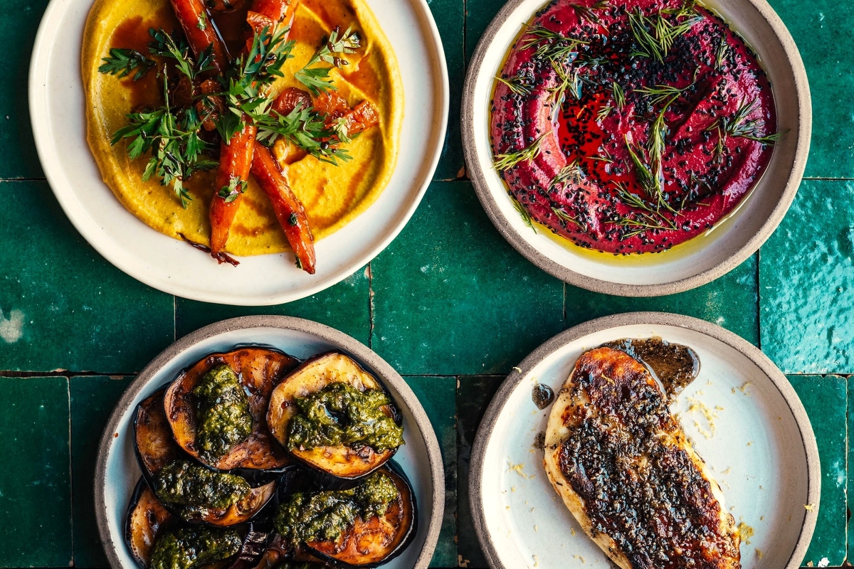 Four colorful Middle Eastern dishes in ceramic bowls on green tiles: roasted carrots with herbs, beetroot dip with rosemary, grilled eggplant with green sauce, and charred flatbread.