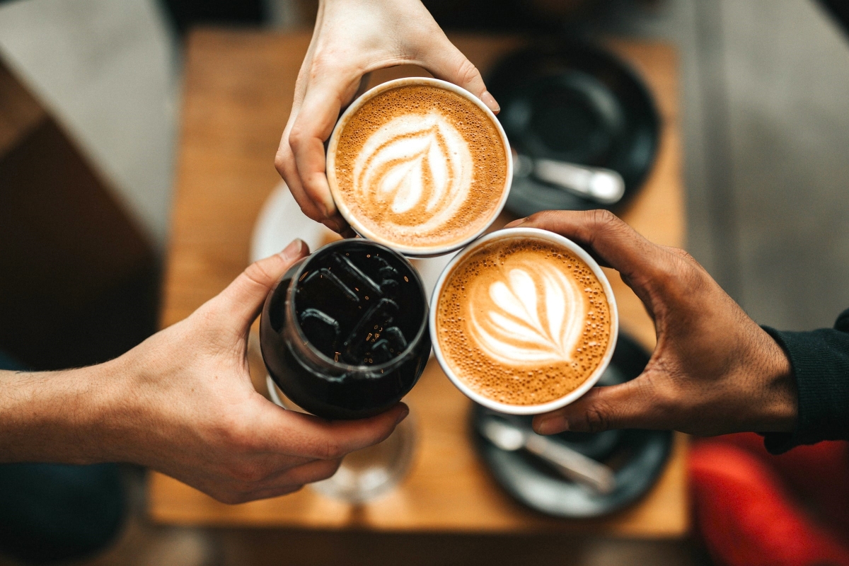 Three hands holding coffee cups together - two lattes with leaf latte art and one black coffee - viewed from above.