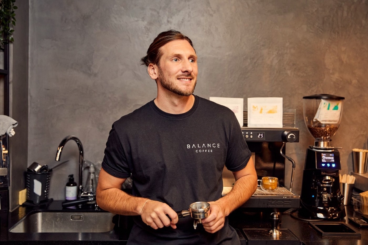A smiling barista in a black Balance Coffee t-shirt stands behind an espresso machine holding a portafilter in a modern coffee shop.