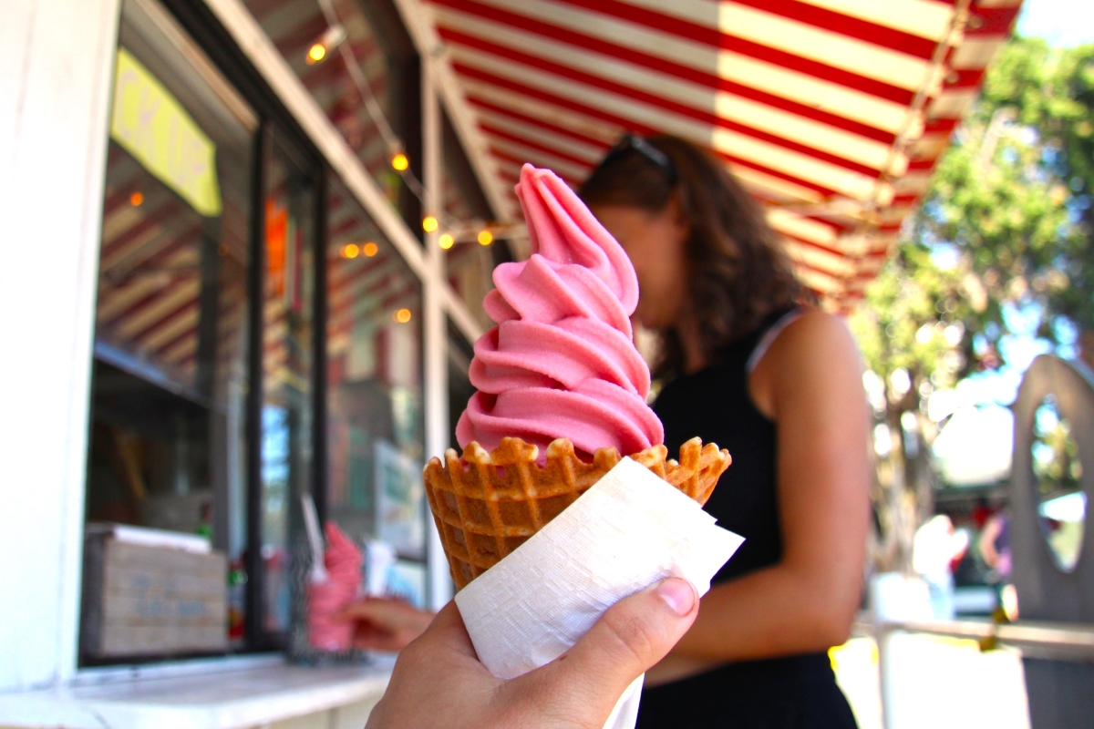 A hand holds a pink soft serve ice cream in a waffle cone on a sunny street with red and white striped awning.