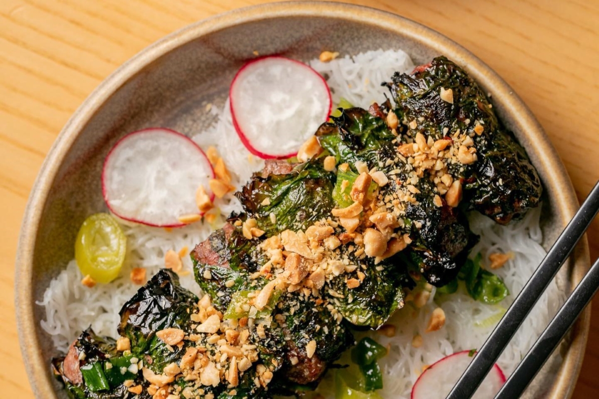 A bowl of rice noodles topped with charred greens, crushed peanuts, and sliced radishes, served with chopsticks.