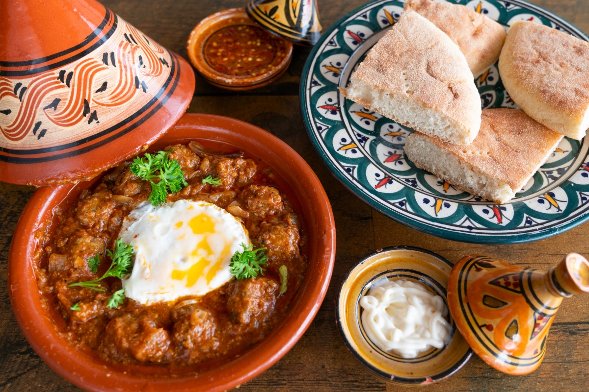 Moroccan tagine with meatballs and fried egg served alongside traditional bread and harissa sauce on colorful ceramic dishes.