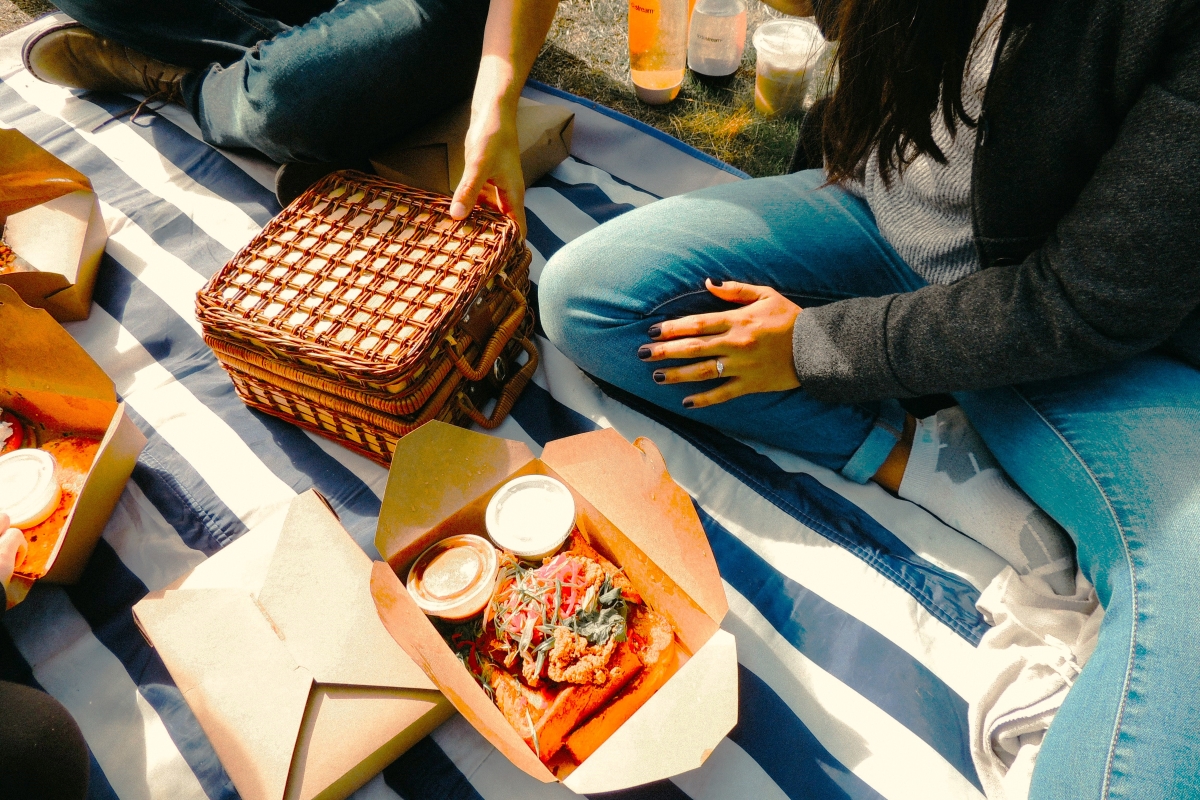 People enjoying takeaway food from cardboard containers on a striped picnic blanket in a sunny park setting.
