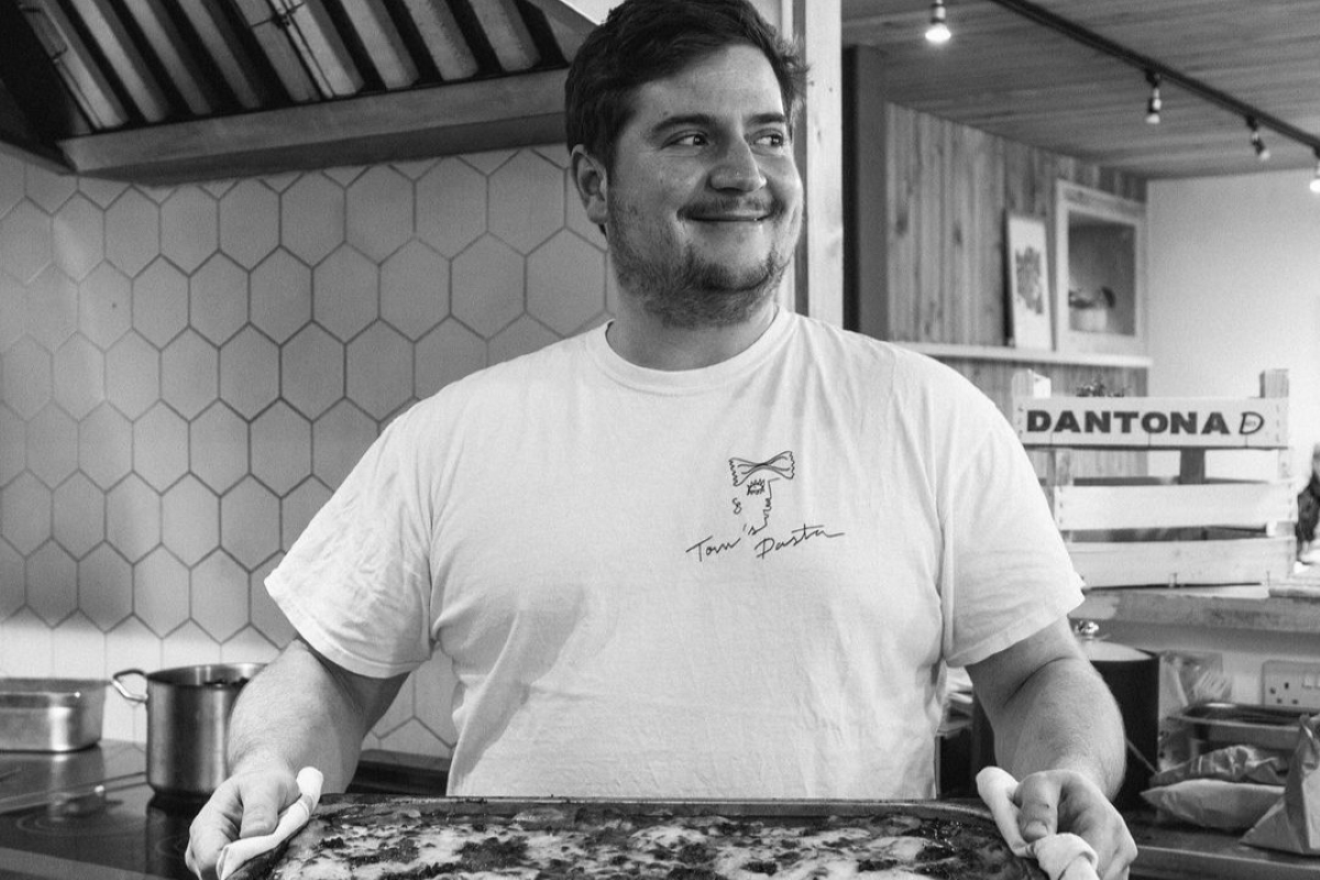 A smiling chef in a white t-shirt holds a freshly baked pizza in a modern kitchen with hexagonal tile backsplash.