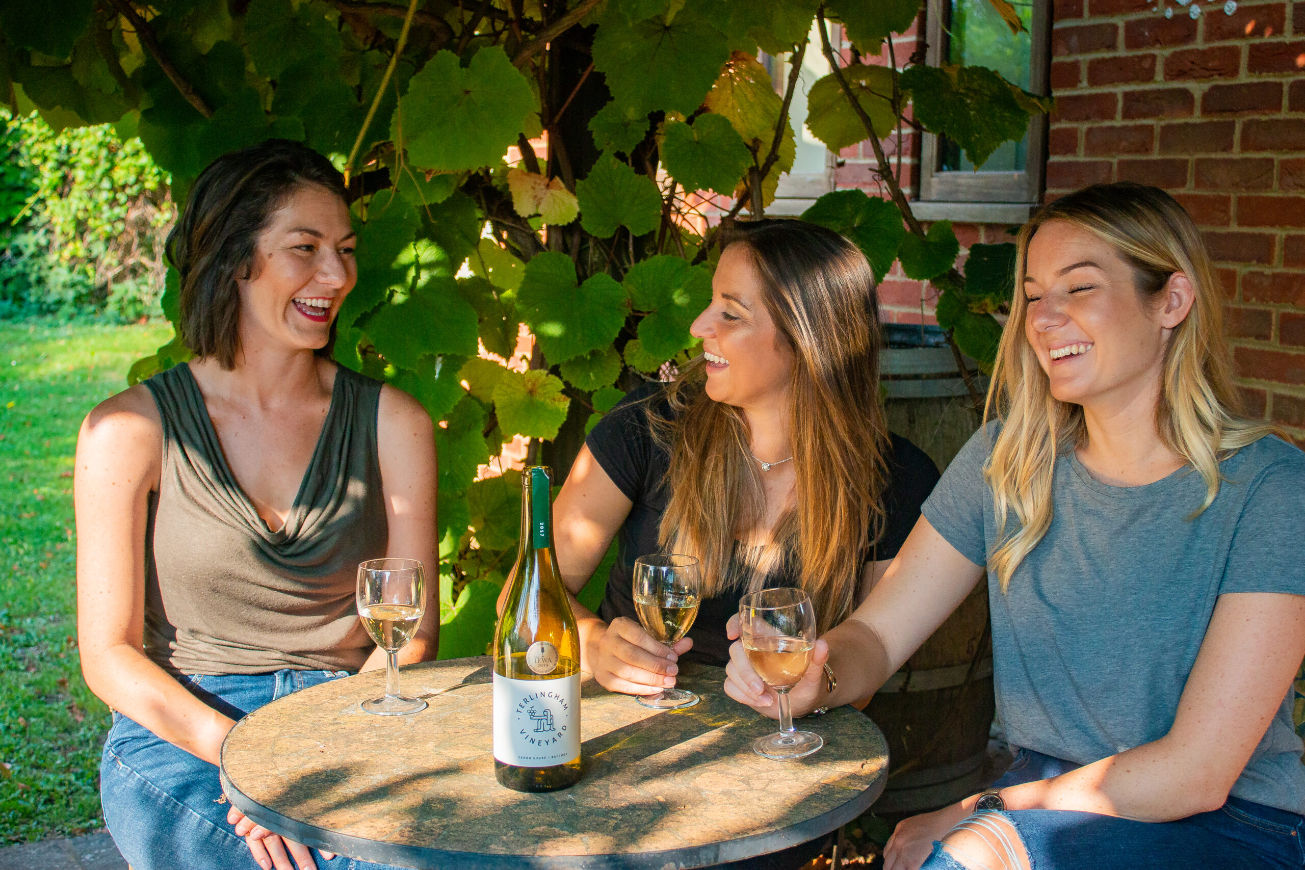 Three women laughing and enjoying white wine at an outdoor table under grapevines at a vineyard.