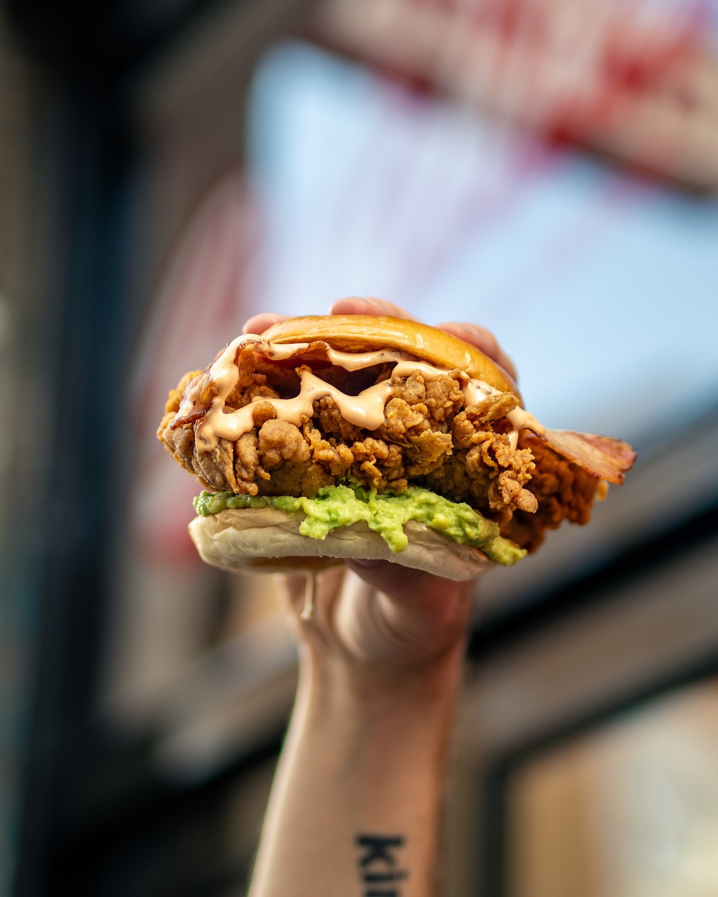 Hand holding a loaded fried chicken sandwich with avocado, cheese sauce, and crispy coating on a sesame bun.