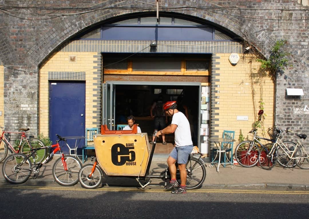 A cyclist with a cargo bike stops outside e5 Bakehouse, located in a brick archway with bicycles parked nearby.