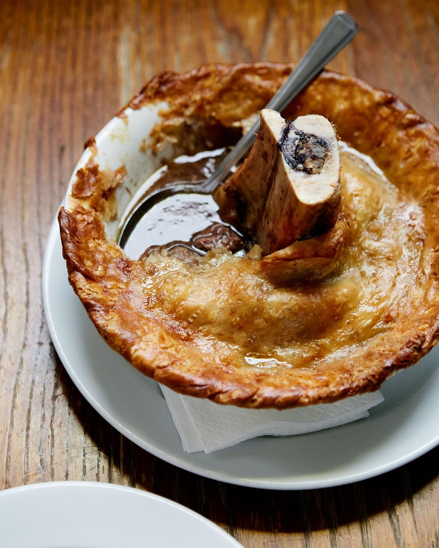 A golden Yorkshire pudding filled with rich gravy and topped with roasted bone marrow, served on a white plate with a spoon.