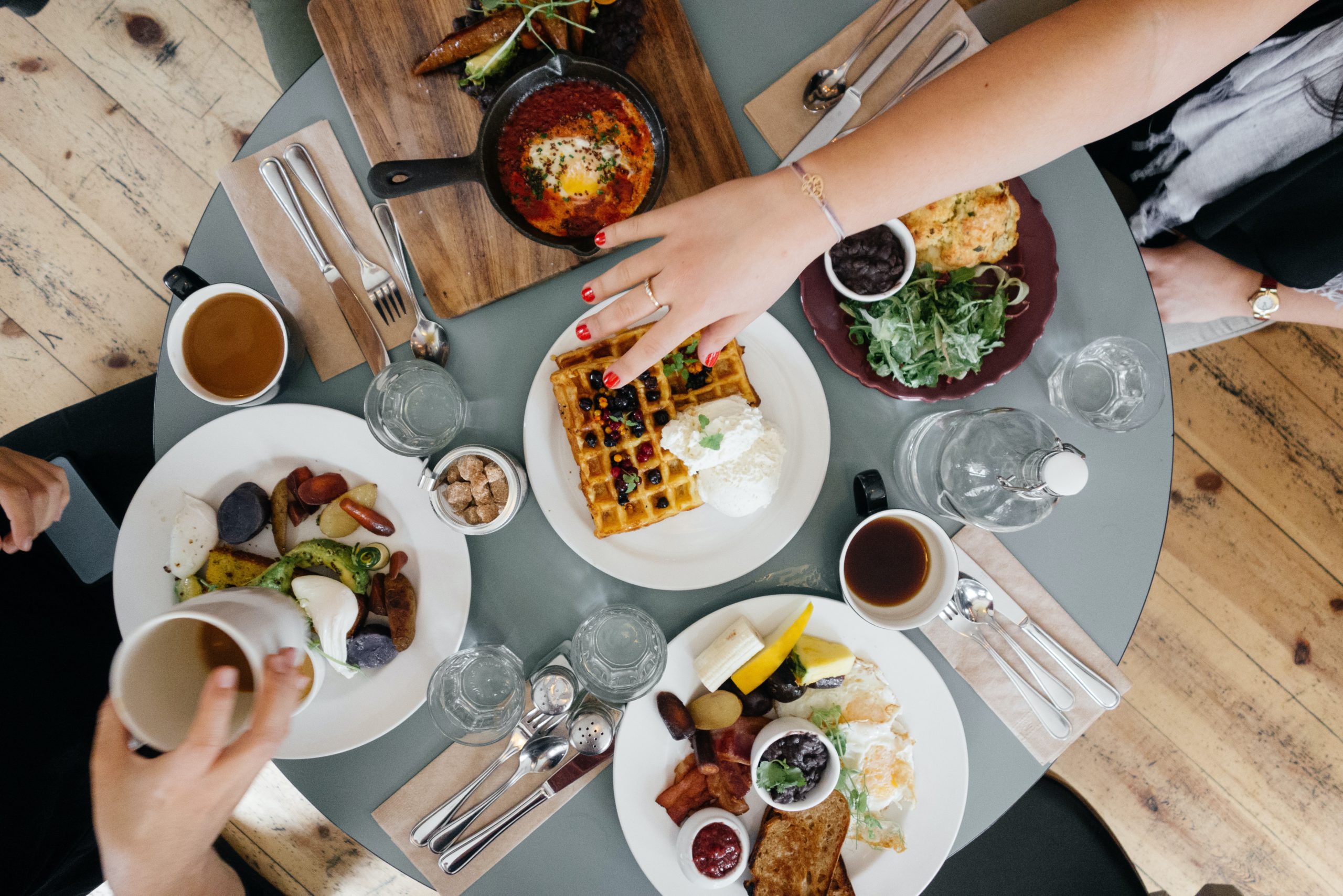 Overhead view of a brunch table with waffles, eggs, bacon, shakshuka in a cast iron pan, and coffee cups shared between diners.