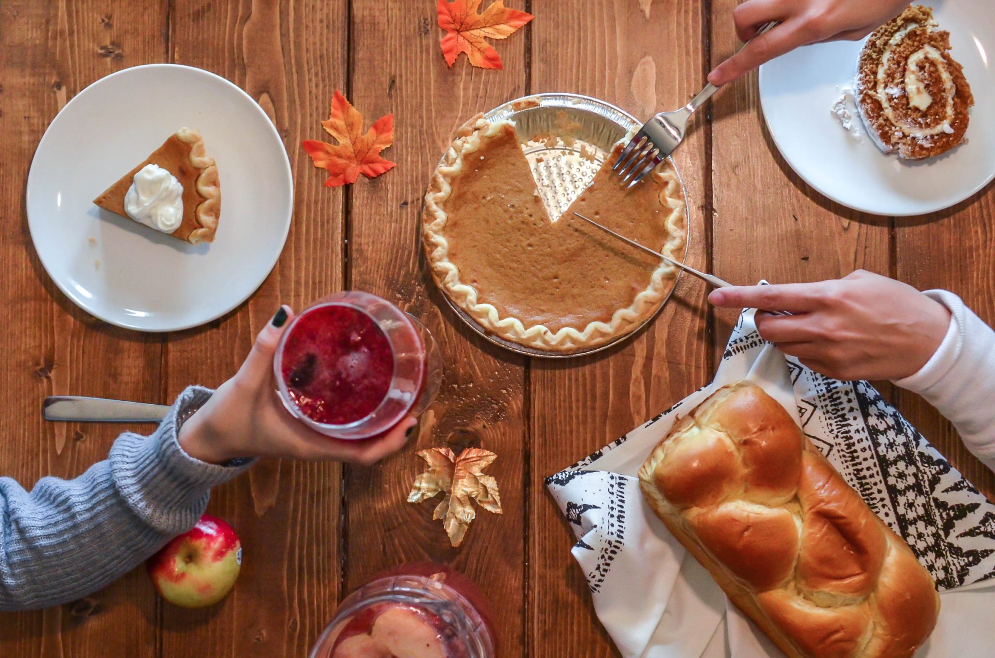 Overhead view of Thanksgiving feast with pumpkin pie, cranberry sauce, challah bread, and autumn leaves on wooden table.