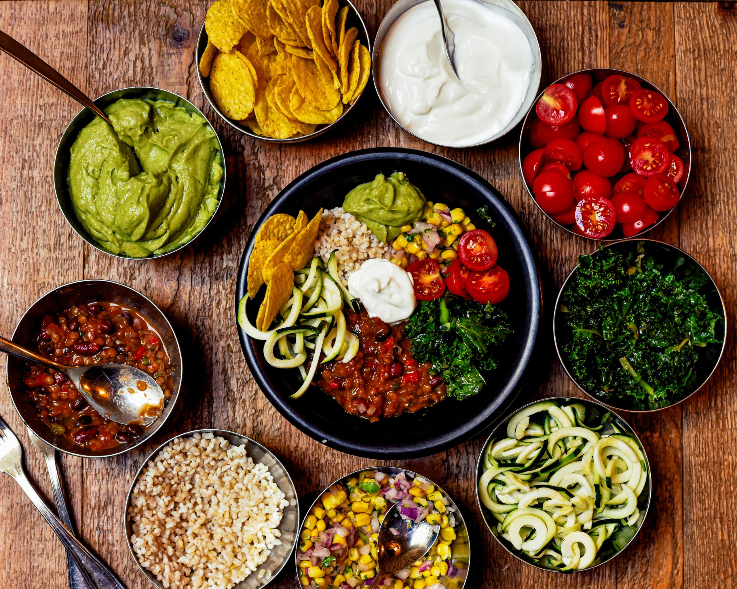 Colorful vegan bowls filled with guacamole, salsa, quinoa, cherry tomatoes, corn salad, kale, and zucchini noodles arranged on wood.