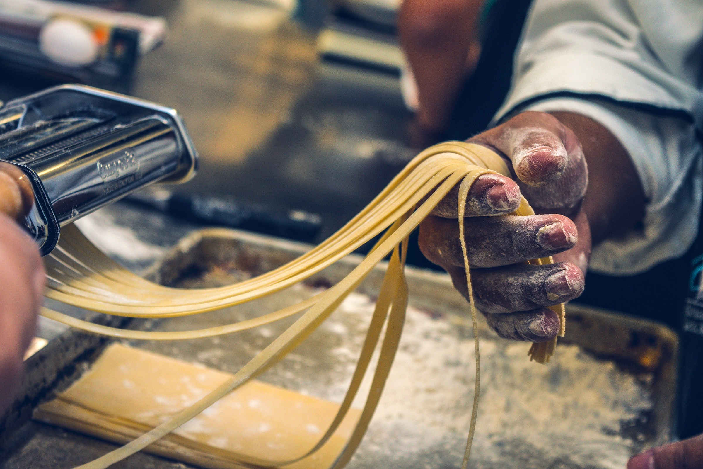 Hands using a pasta machine to roll out fresh yellow linguine noodles on a floured wooden surface.