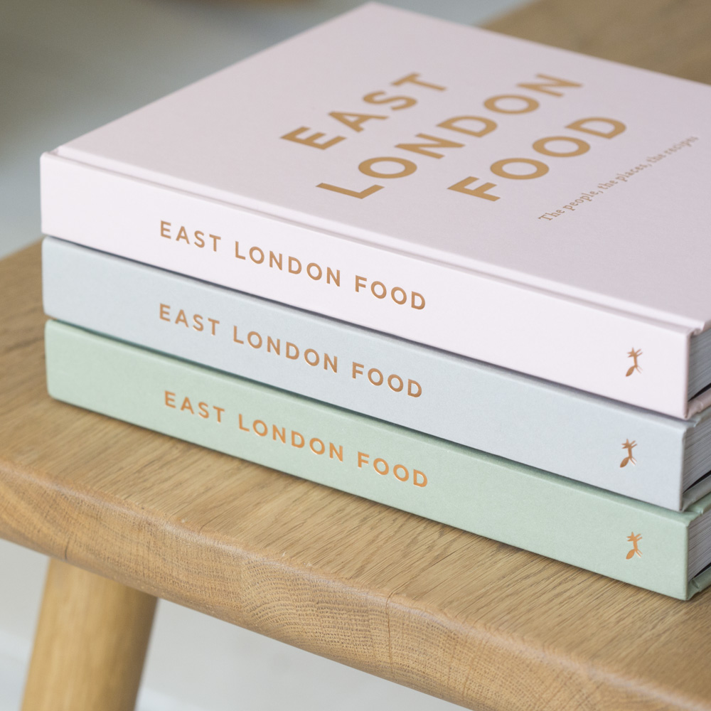Three stacked copies of the "East London Food" cookbook in pink, white, and mint green covers on a wooden stool.