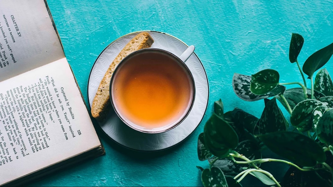 A cup of amber tea with a biscotti on a white saucer next to an open book and green plant on a turquoise wooden surface.