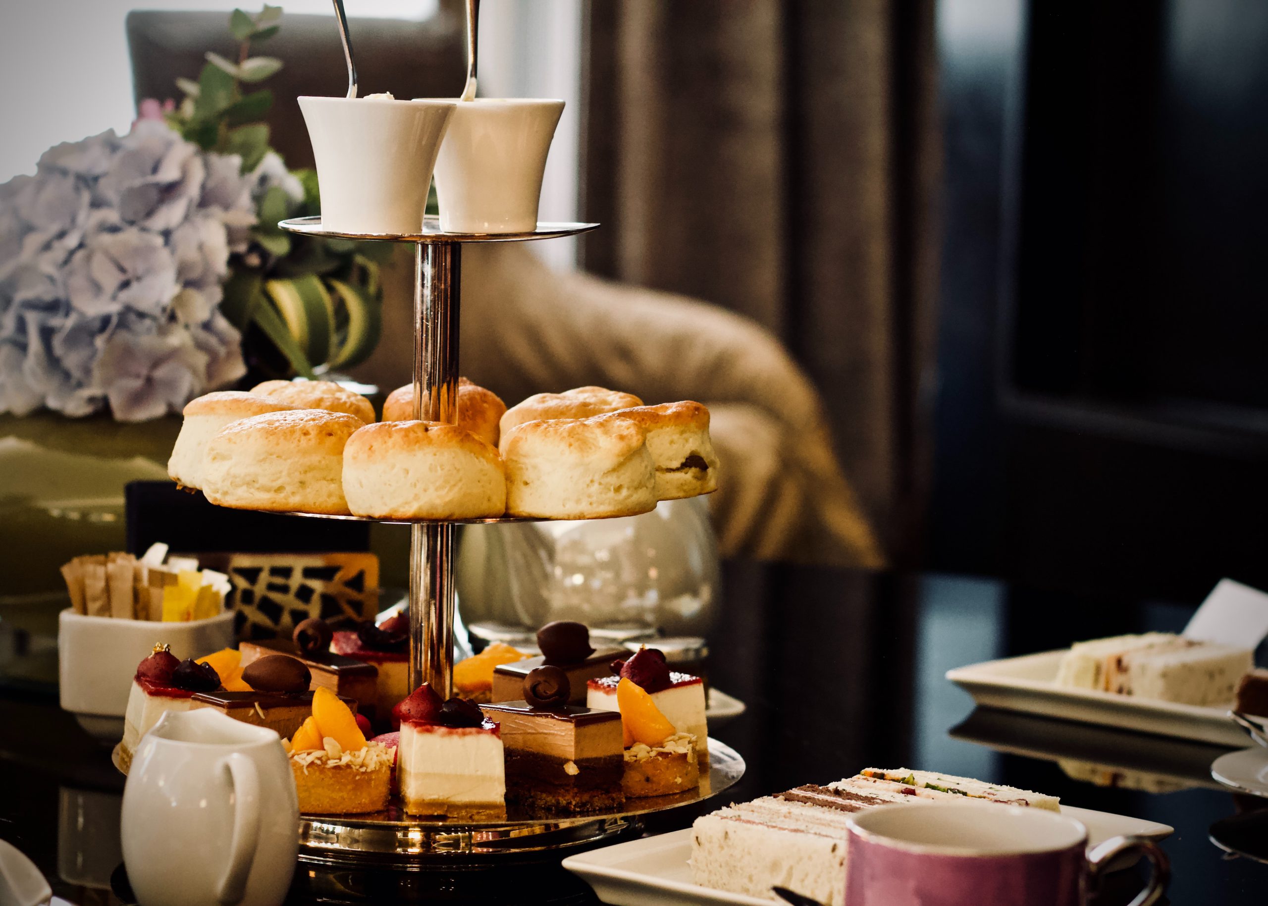 An elegant three-tiered stand displays scones on the middle tier and colorful petit fours on the bottom, with teacups and flowers nearby.