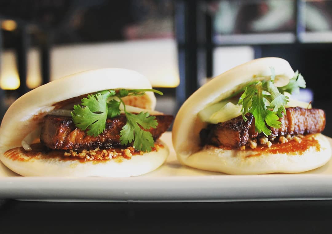 Two fluffy white bao buns filled with glazed pork belly and fresh cilantro on a white plate.