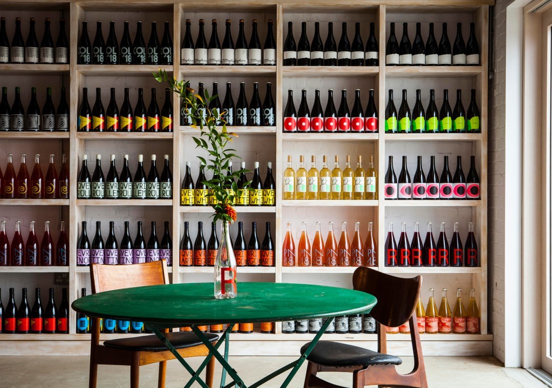 A wine shop interior with wooden shelves displaying colorful wine bottles and a green bistro table with two chairs.