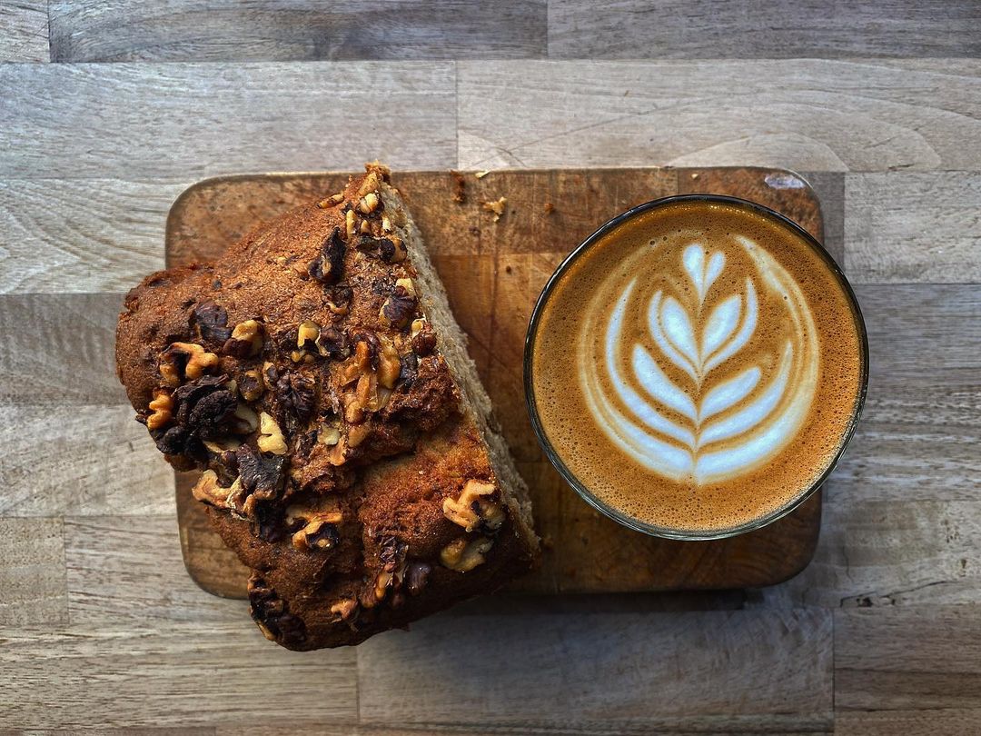 A latte with leaf-pattern foam art and chocolate walnut bread slices on a wooden board, photographed from above.