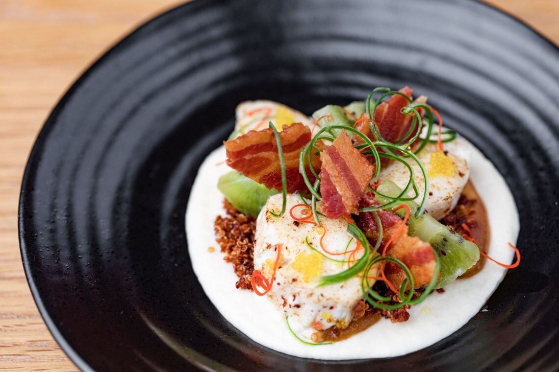 A gourmet dish with hard-boiled eggs, crispy bacon, and colorful garnishes artfully plated on a white dish in a black bowl.