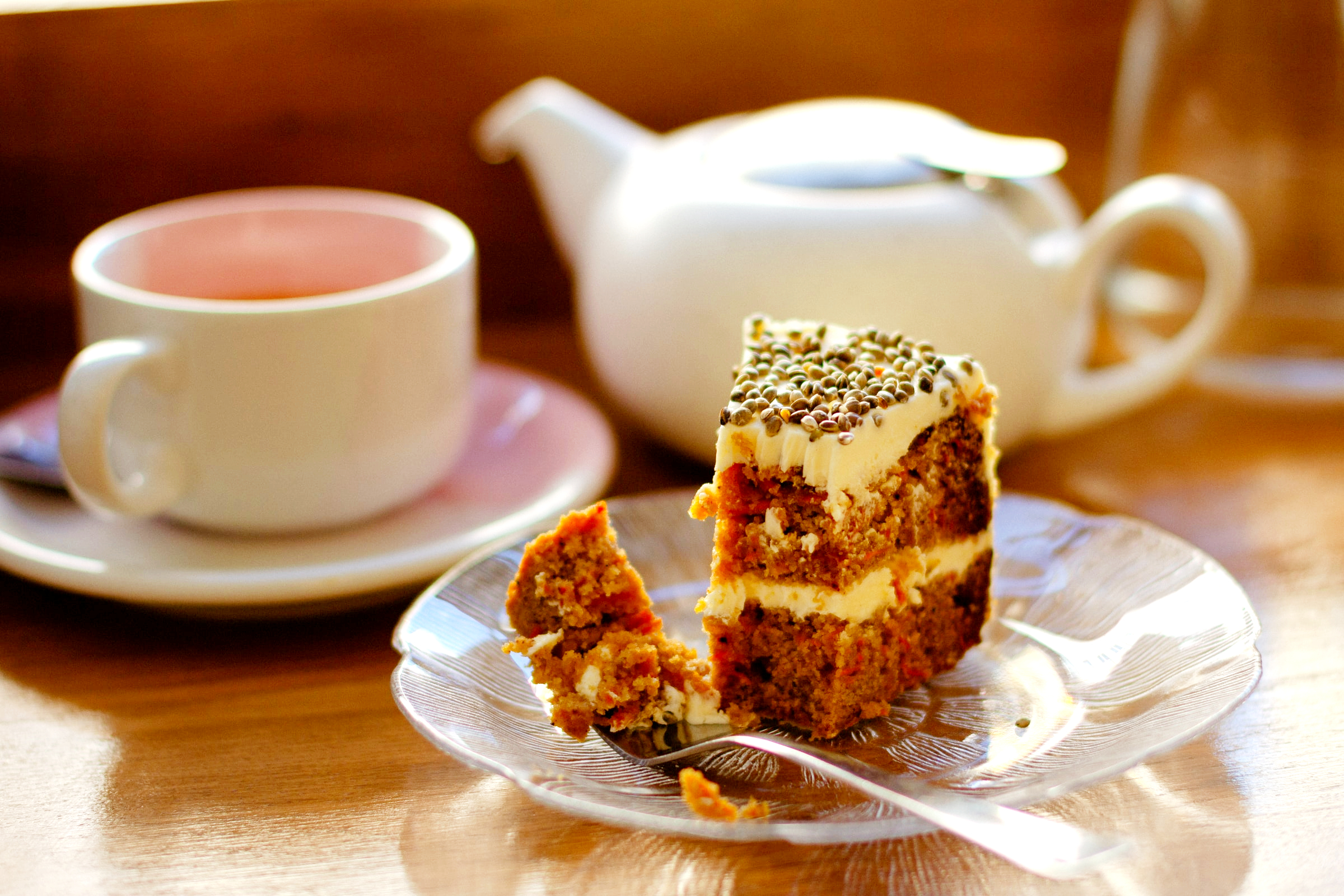 A slice of layered carrot cake with cream cheese frosting sits on a glass plate beside a white teacup and teapot.