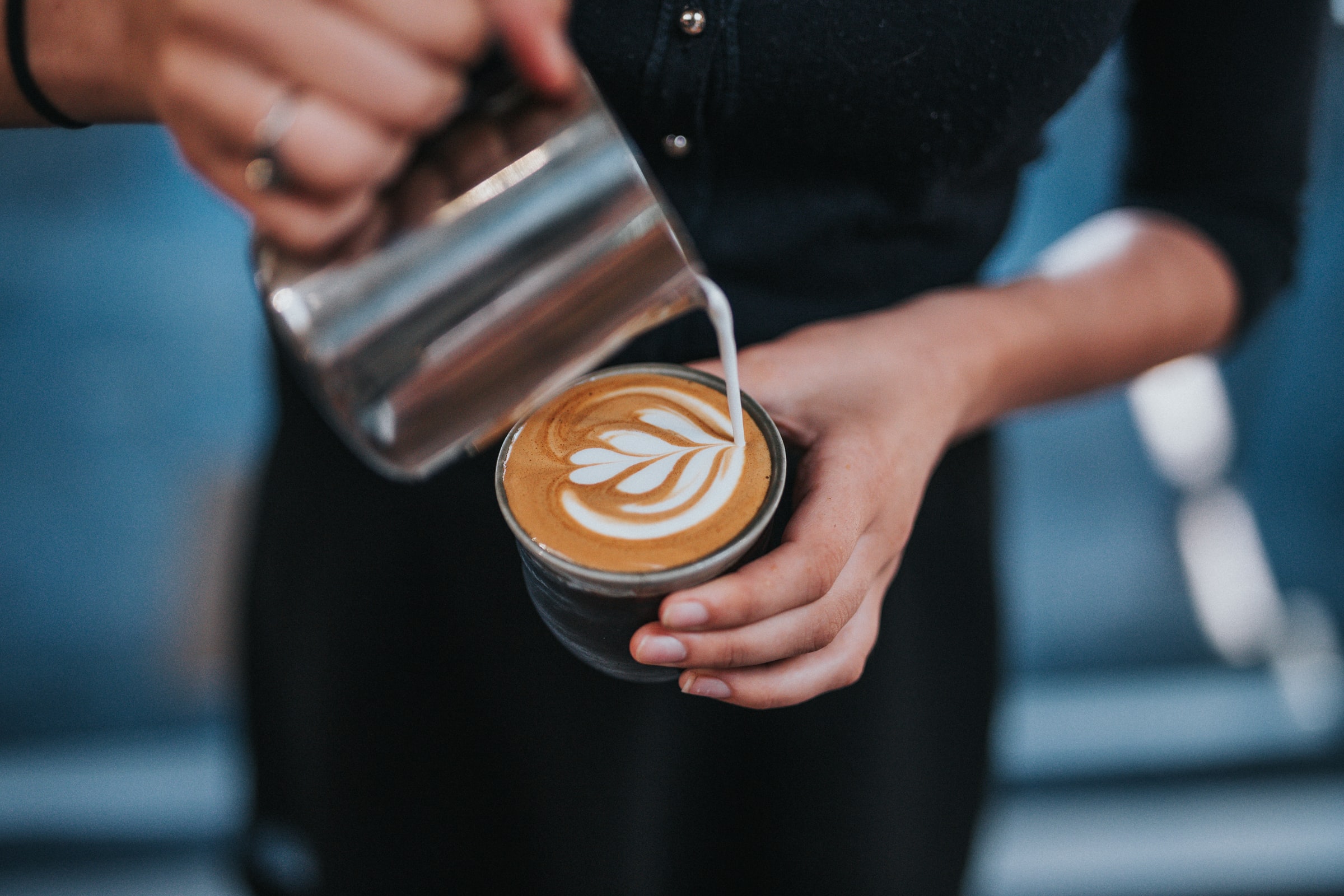 A barista pours steamed milk from a metal pitcher into a coffee cup, creating latte art with a leaf design.