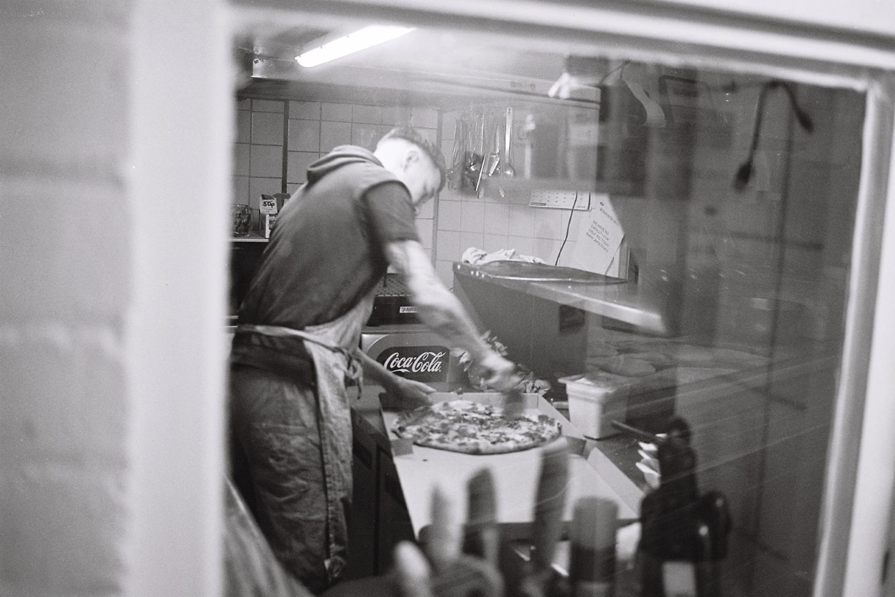 A pizza chef in white uniform and cap works in a kitchen, preparing pizza at the counter with a Coca-Cola cooler nearby.